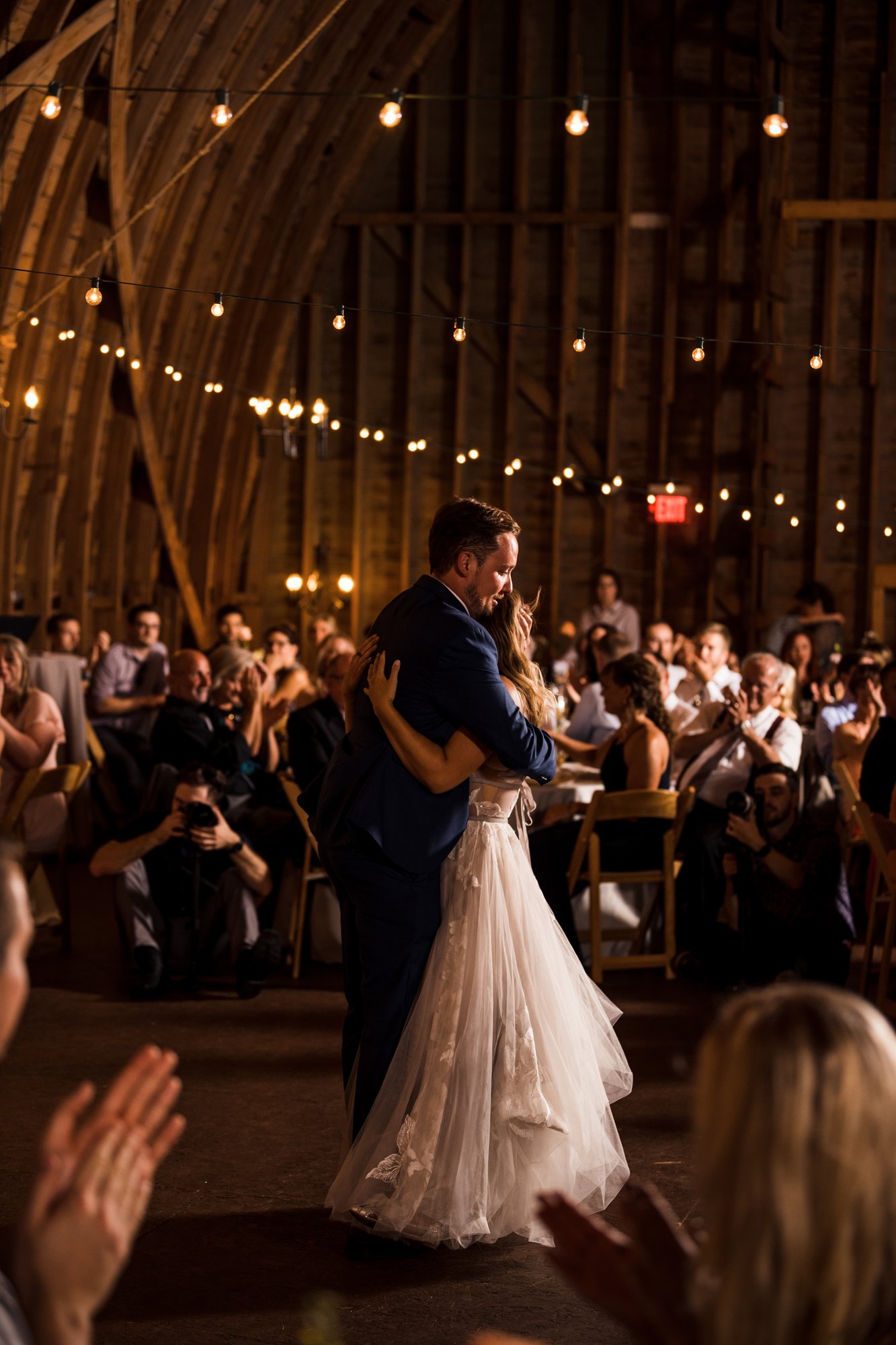 A bride walks the aisle inside the barn beneath strings of warm light.