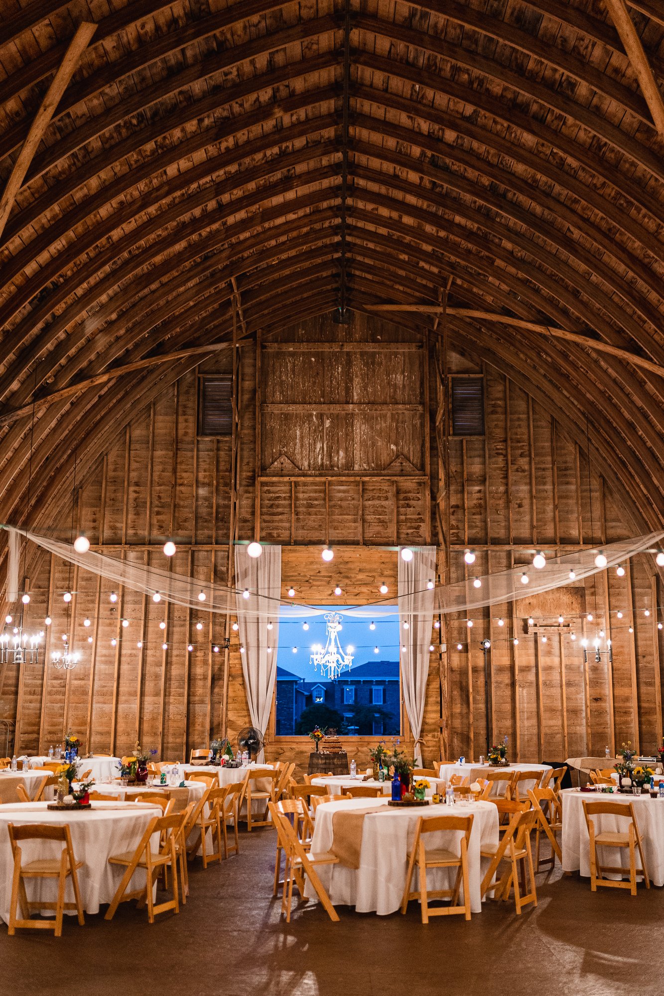 The full length of the barn interior with round tables set beneath the arched timber ceiling and string lights.