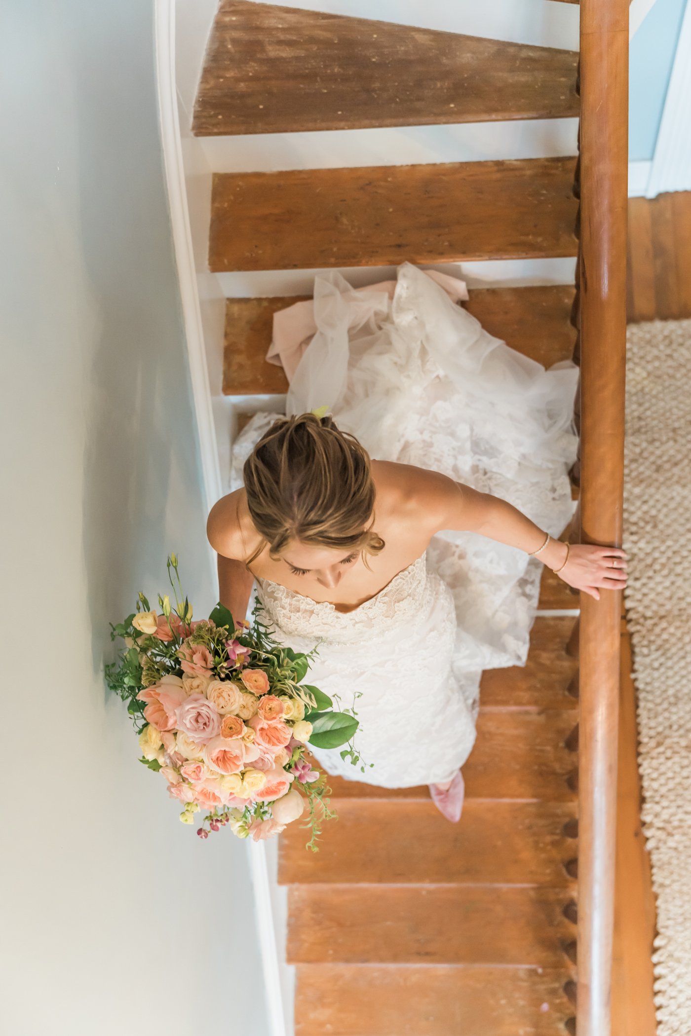 A bride carries her peach bouquet down the farmhouse staircase.