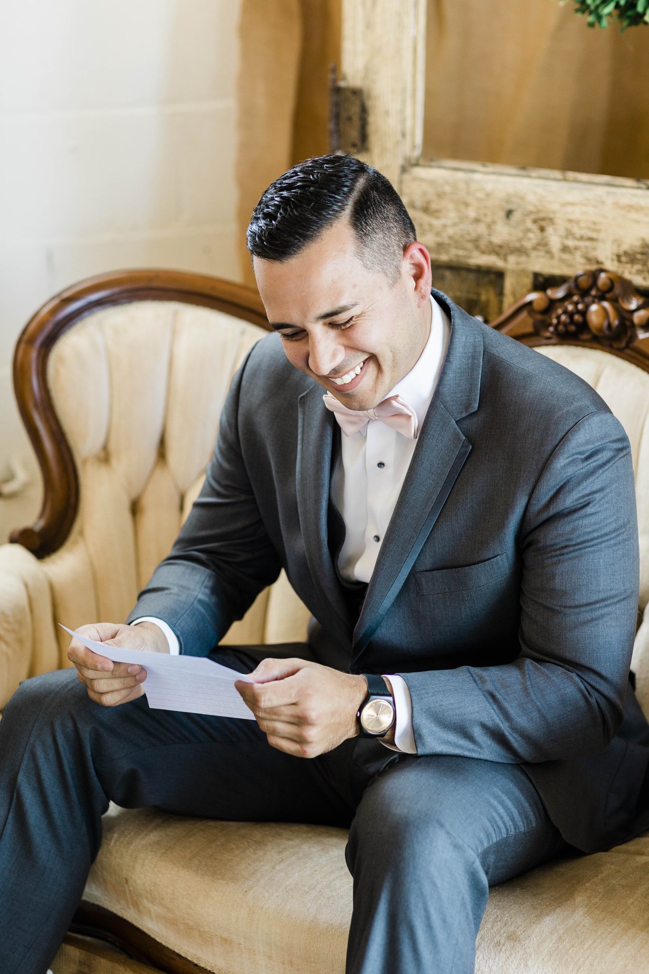 The groom sits on a settee in the farmhouse reading a letter before the ceremony.