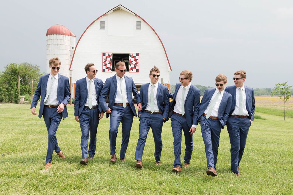 Seven groomsmen in blue suits walking the field in front of the white barn.
