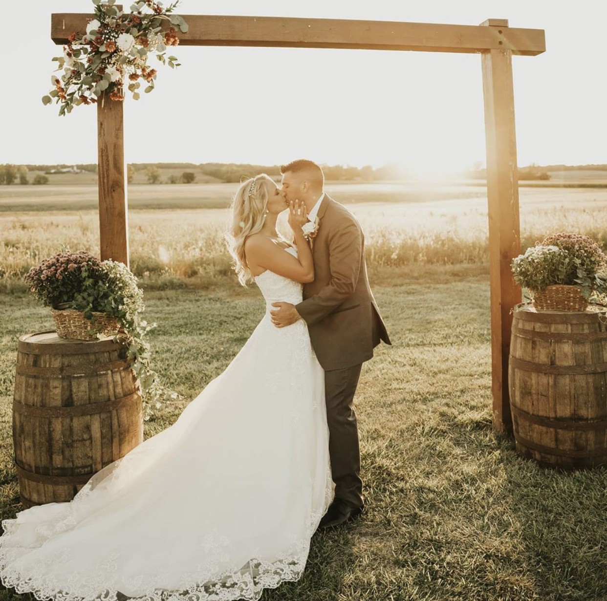 A couple under the floral arch between two whiskey barrels at golden hour.