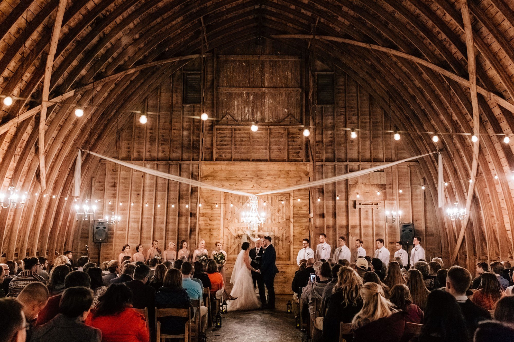 A full ceremony inside the barn, guests filling the seats beneath the chandelier and string lights.