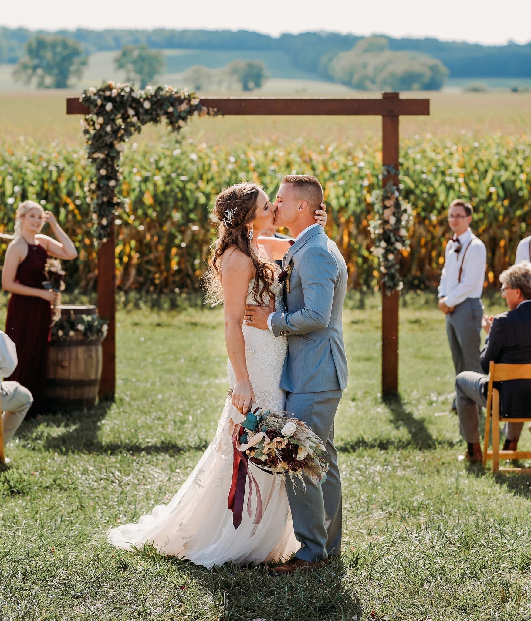 A couple exchanging vows under a floral arch with the cornfield behind them.