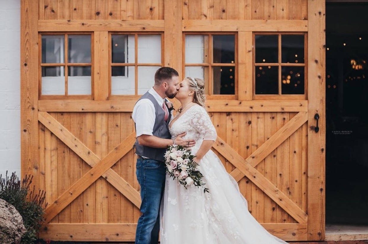 A couple kissing in front of the tall wooden barn doors.