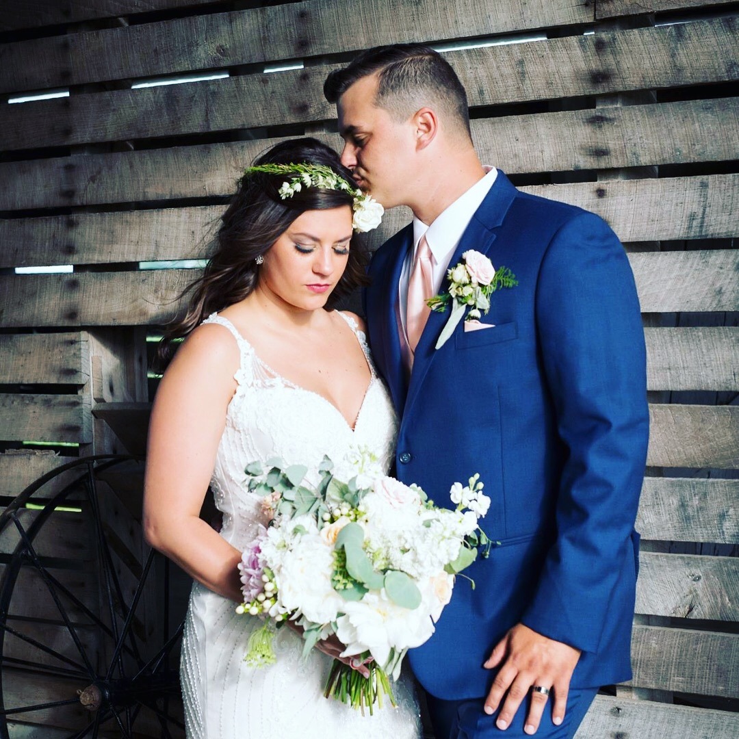 A couple close together against the dark barn siding, greenery in her hair.