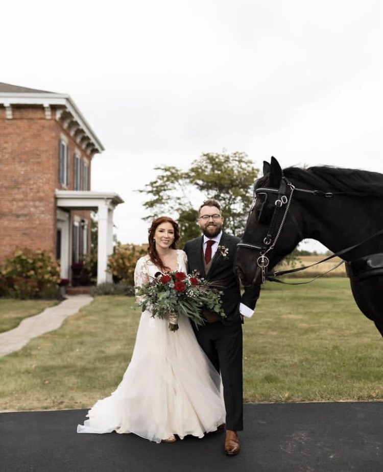 A couple standing with a black horse on the drive beside the farmhouse.