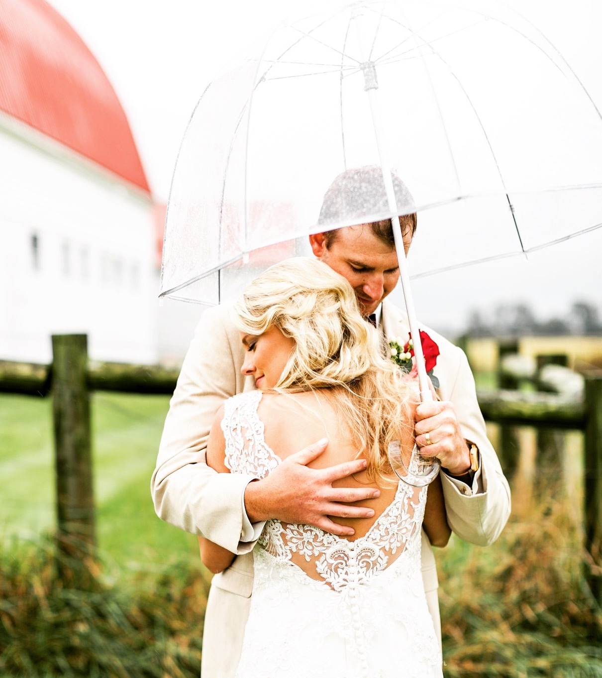 A couple under a clear umbrella in the rain outside the barn.