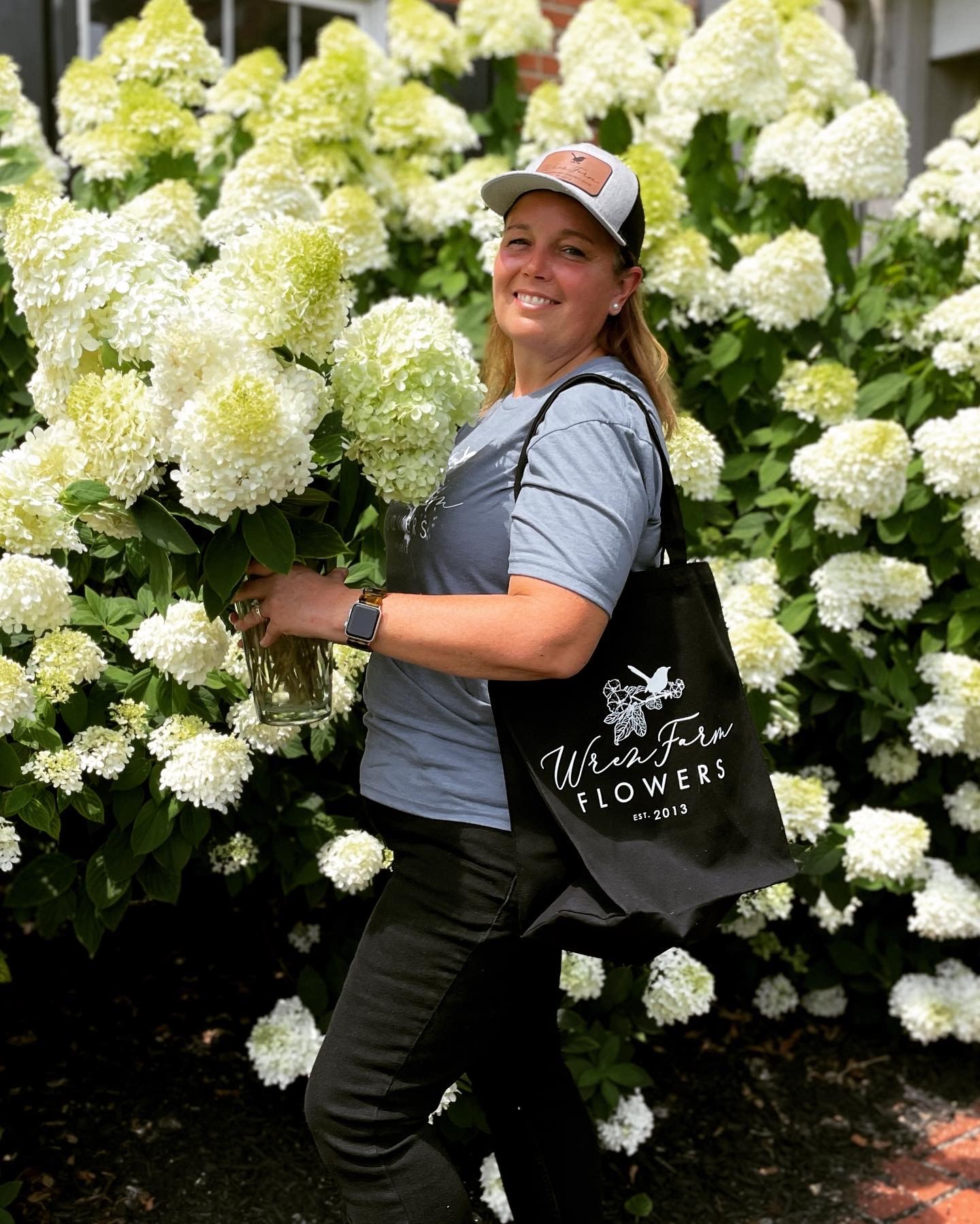 Hydrangeas gathered into a vase in front of the blooming bushes, a Wren Farm Flowers tote on the shoulder