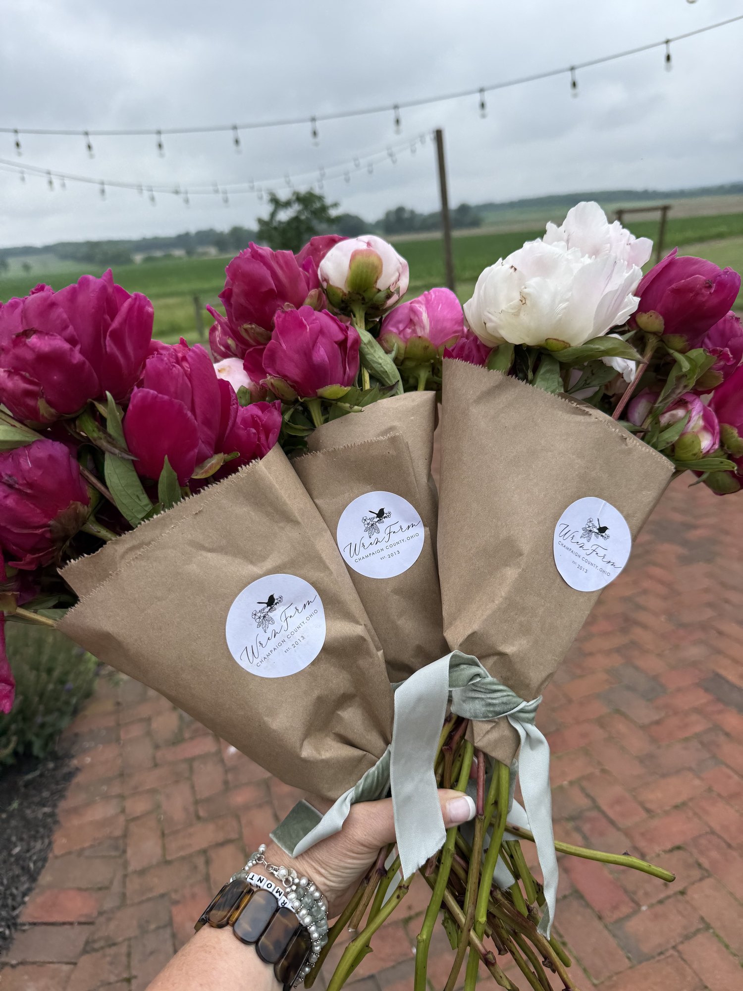 Three peony bouquets wrapped in kraft paper with Wren Farm stickers and sage ribbon, held over the brick patio
