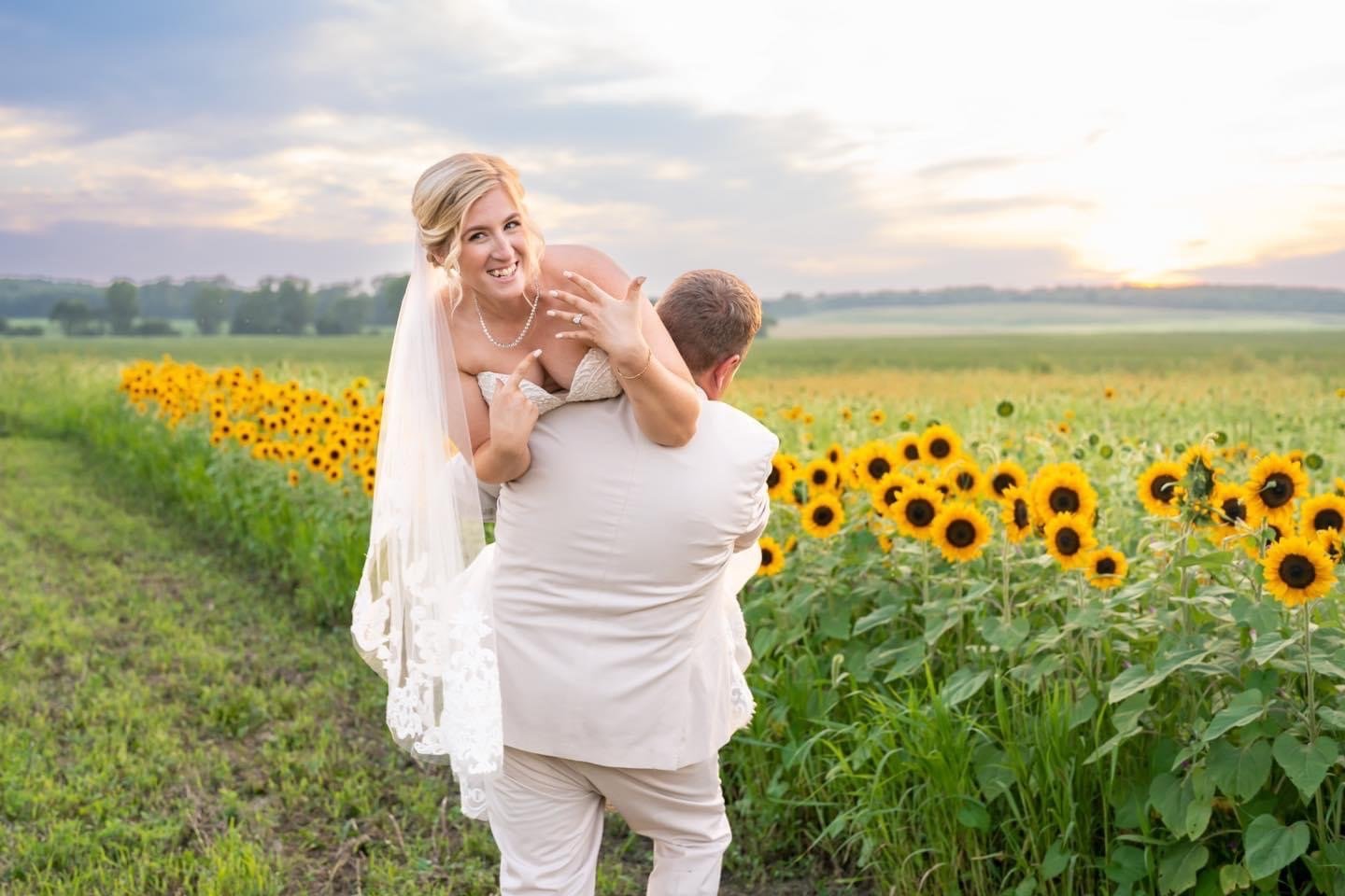 A groom lifts the bride along the edge of the sunflower field at sunset