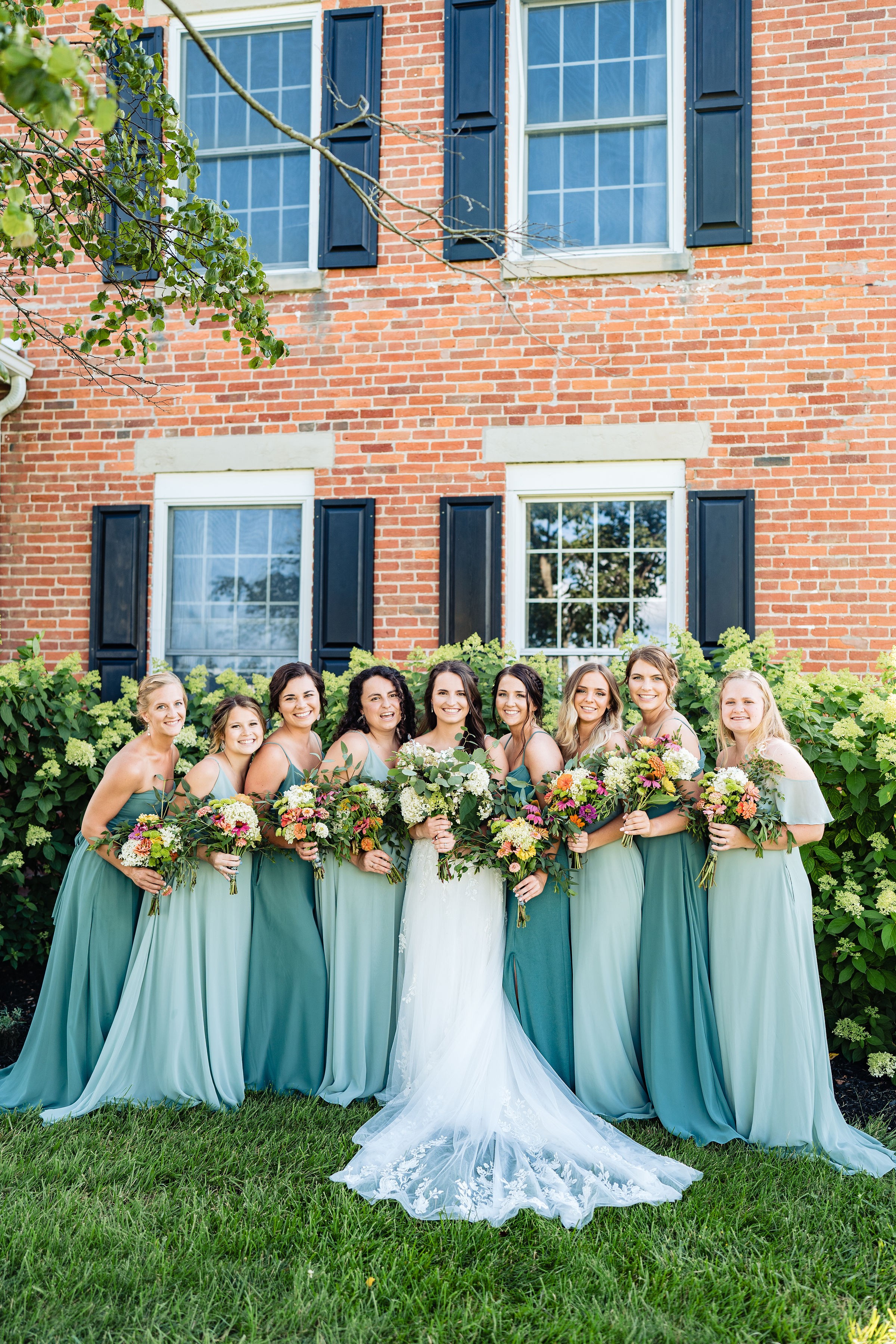 Bridesmaids in teal and sage gathered in front of the brick farmhouse.
