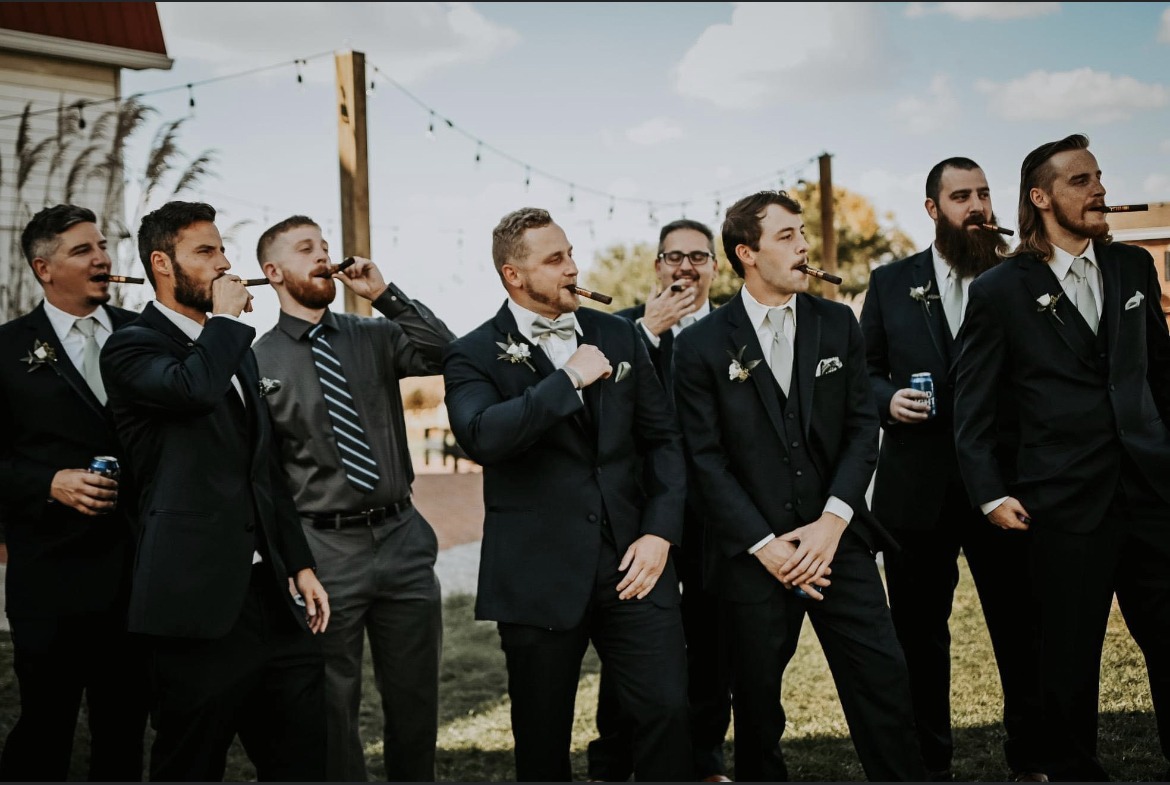 Groomsmen in black tuxedos laughing together outside the barn.
