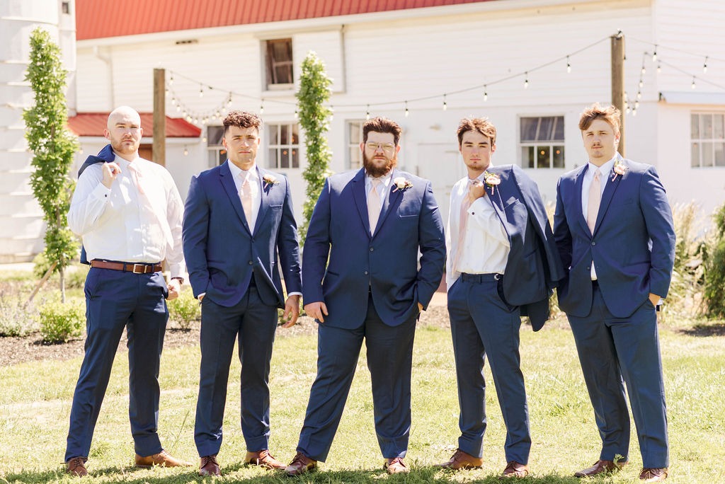 Groomsmen in navy suits in front of the brick farmhouse and its hydrangeas.