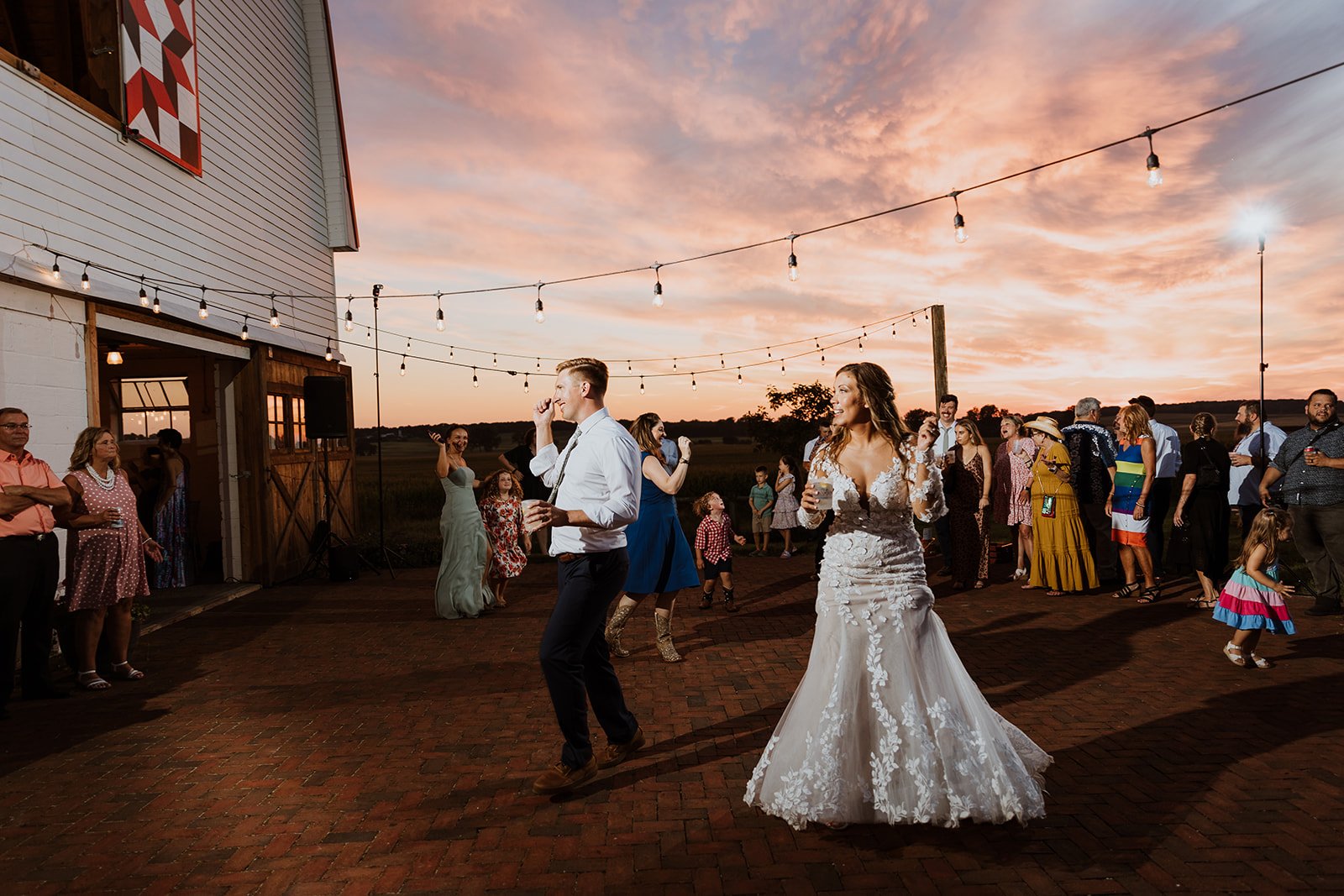 Guests and the newlyweds dance together on the brick patio beneath cafe lights as the sky turns pink.