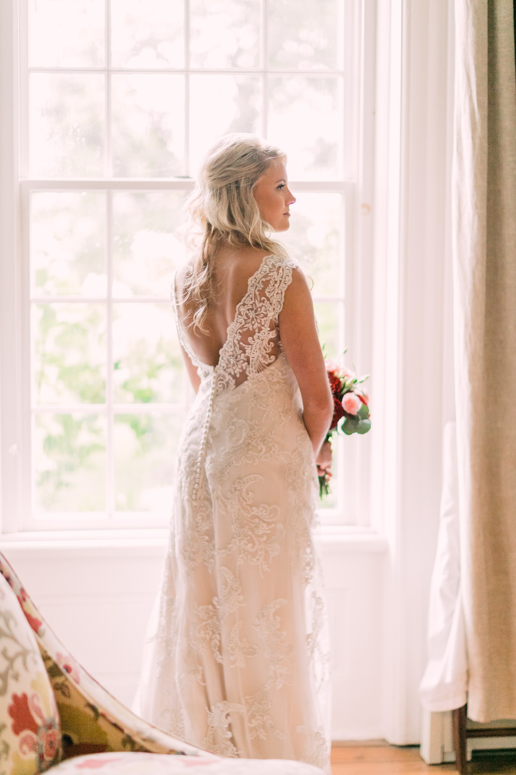 A bride stands beside a bright farmhouse window in her wedding gown.