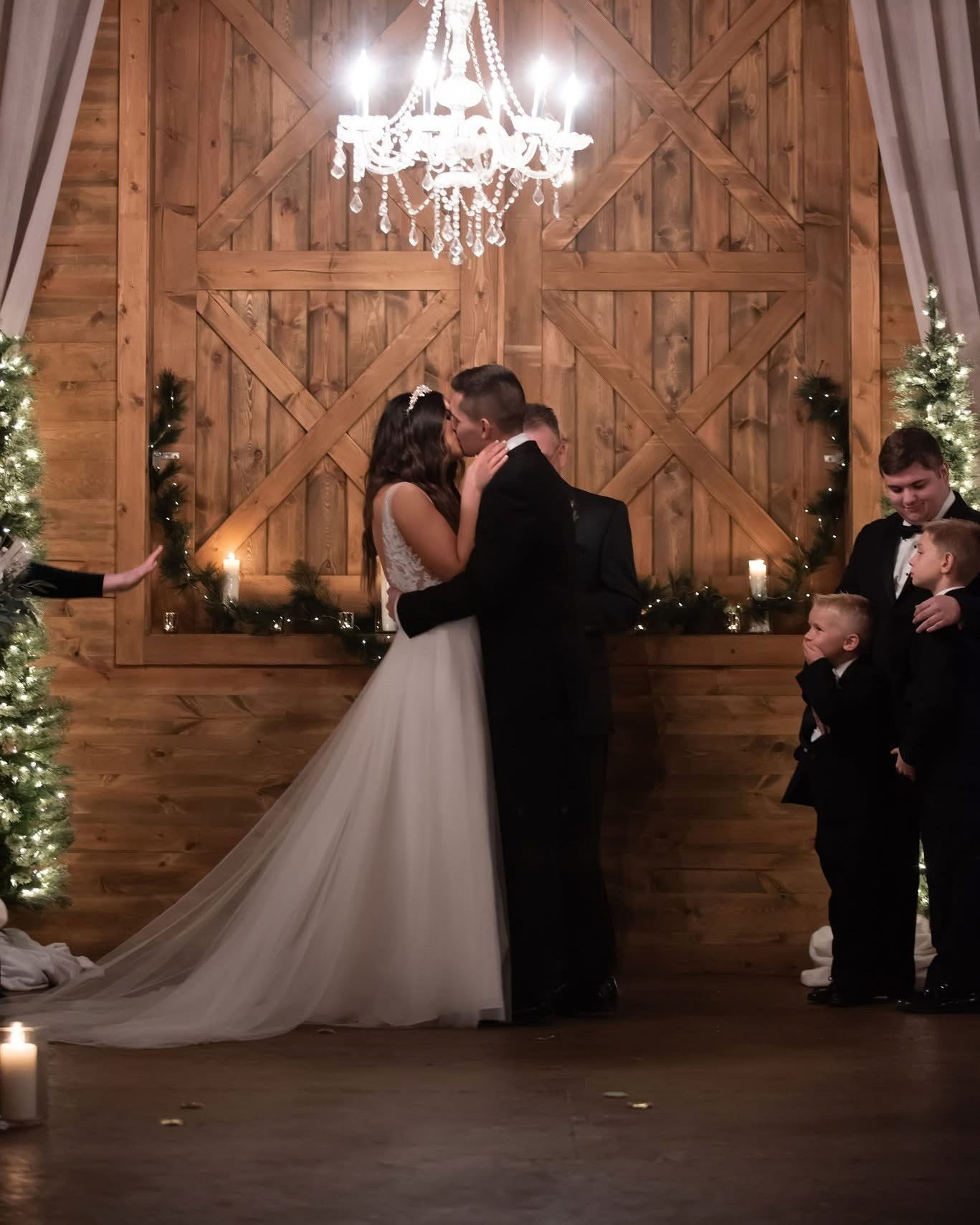 A couple shares their first kiss inside the barn beneath a crystal chandelier, with candles and greenery along the wall behind them.