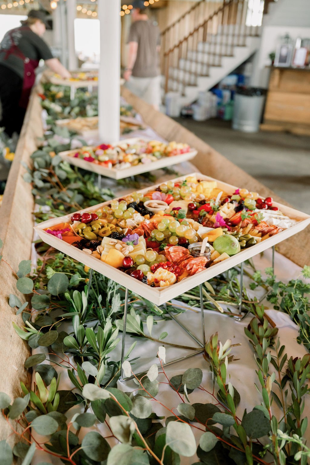 Trays of cheese, fruit, and charcuterie set along a greenery-lined table for cocktail hour.