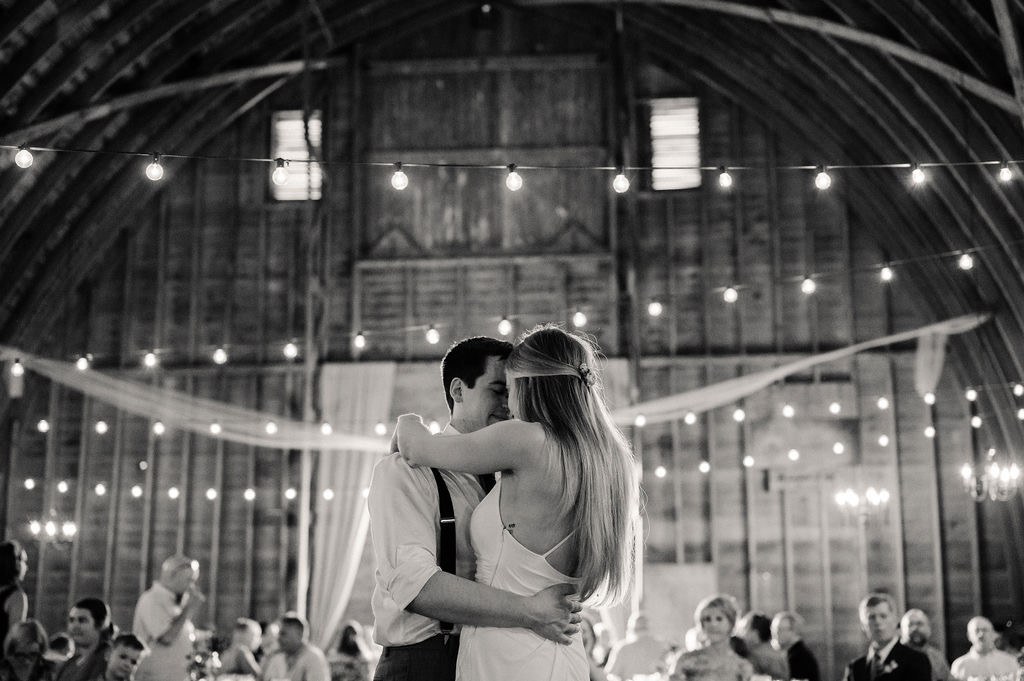 A couple holds each other during their first dance beneath the barn lights.