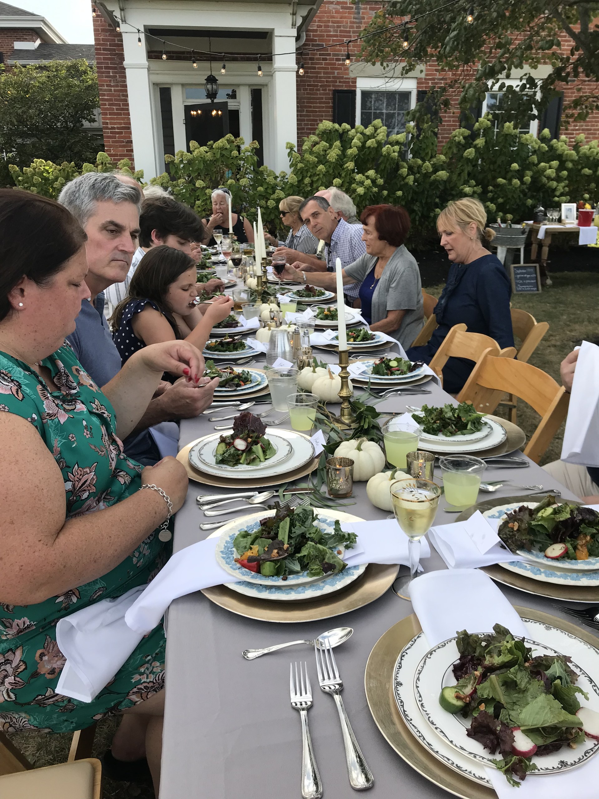 Guests share a long outdoor dinner table in front of the Wren Farm farmhouse.