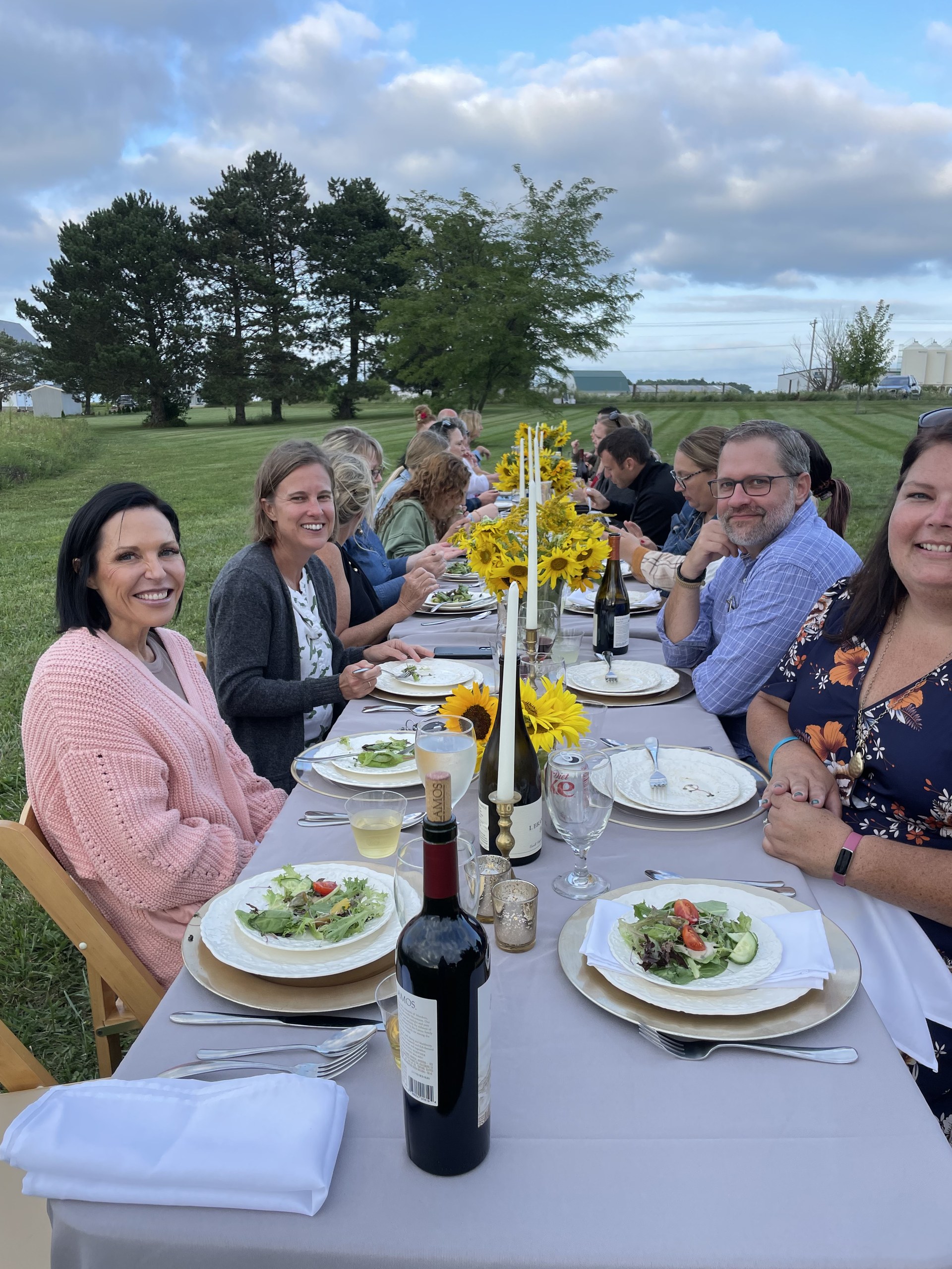 Guests gather around a long table set with sunflowers beside the open field.